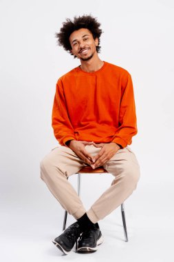 Afro saçlı genç adam gülüyor, beyaz stüdyo geçmişi olan bir sandalyede oturuyor.