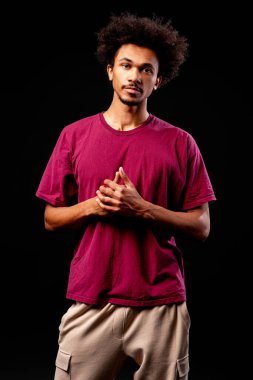 Siyah arka planda kahverengi tişört ve bej kargo pantolon giyen Afro saç modelli genç adam.