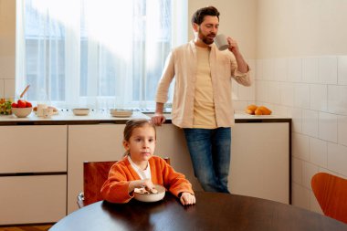 Küçük kız masada mısır gevreği yerken babam ayakta kahve içiyor.