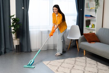 Eldiven giyen, oturma odasını paspaslayan, temizlik yapan genç bir kadın.