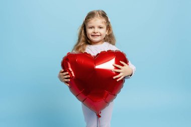 Gülümseyen genç bir kız, elinde kırmızı kalp şeklinde bir balonla mavi bir stüdyonun arka planında