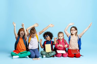 Çeşitli ilkokul çocukları ellerinde okul ekipmanlarıyla yerde oturuyor, el kaldırıyorlar, öğrenmeye hazırlar.