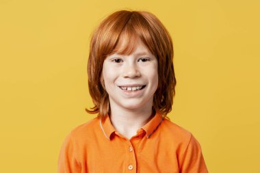 Çilli kızıl saçlı çocuk, parlak sarı bir arka plana rağmen kameraya neşeyle gülümsüyor.