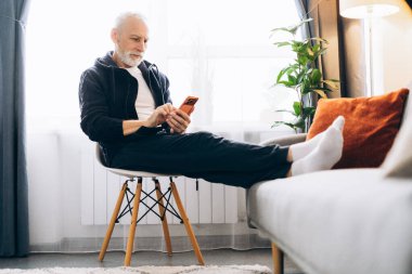 Cep telefonu uygulaması kullanarak cep telefonu tutan kendinden emin, olumlu gülümseyen yaşlı bir adam. Evdeki oturma odasında sandalyede oturan kısa mesaj. Çevrimiçi teknoloji kavramı
