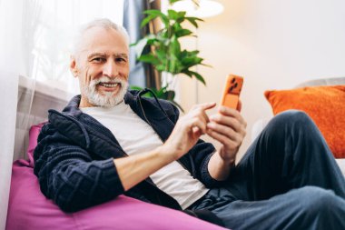 Gülümseyen, mutlu, son sınıf öğrencisi cep telefonu tutan, sohbet eden, mesaj atan rahat oturma odasında oturan kameraya bakan. Sosyal medya kavramı