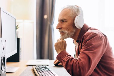 Evde çalışan yaşlı, gri saçlı bir adamın portresi. Bilgisayar kullanıyor, iş yerinde kulaklık takıyor. Uzak görev konsepti