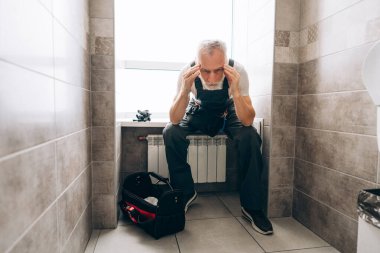 Yaşlı, profesyonel, gri saçlı, iş kıyafetleri giyen, banyoda sifon tamir aletleri kullanan bir adam. Onarım kavramı