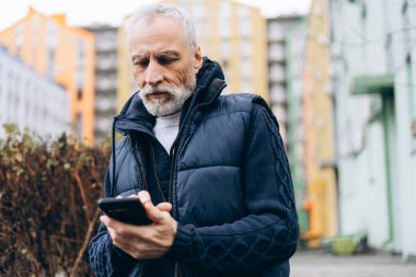 Yakışıklı, ciddi bir kıdemli adam cep telefonu kullanıyor cep telefonu uygulaması kullanıyor, sohbet ediyor, sokakta mesaj yazıyor. Çevrimiçi teknoloji kavramı