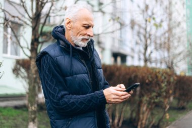 Çekici, ciddi bir kıdemli adam cep telefonu kullanıyor cep telefonu uygulaması kullanıyor, sohbet ediyor, sokakta mesaj yazıyor. Çevrimiçi teknoloji kavramı