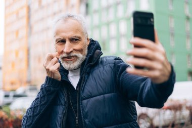 Gülümseyen, gri saçlı, cep telefonu tutan, selfie çeken, blogcu, sokakta video kaydeden son sınıf öğrencisi. Çevrimiçi teknoloji kavramı