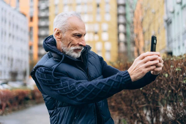 Yakışıklı, gri saçlı, cep telefonu tutan, fotoğraf çeken, blogcu, sokakta video kaydeden yaşlı bir adam. Çevrimiçi teknoloji kavramı