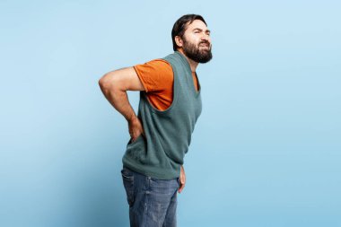 Üzgün, sakallı adamın sırt ağrısı var. Mavi arka planda tek başına duruyor. Spazm, sağlık hizmeti kavramı