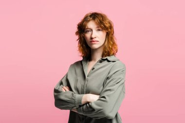 Çapraz kollu, sıradan kıyafetler giymiş, pembe arka planda izole edilmiş kameraya bakan ciddi düşünceli genç bir kızıl kadın. Reklam kavramı