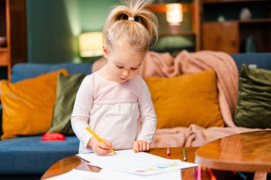 Yaratıcı bir aktiviteden hoşlanan küçük bir kız, oturma odasındaki bir masada çizim yapmaya odaklanmış.