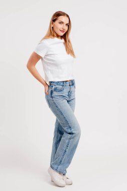 Beyaz gömlekli ve beyaz arka planda kot pantolonlu sıradan poz veren genç bir kadın.