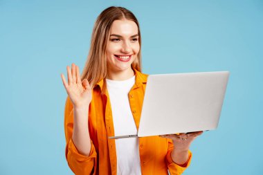 Genç bir kadın gülümsüyor, video aracılığıyla birini selamlıyor, dizüstü bilgisayarını mavi arka planda tutuyor.