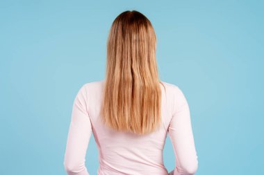 Sırtı kameraya dönük duran, mavi arka planda uzun, sağlıklı sarı saçları olan bir kadın.