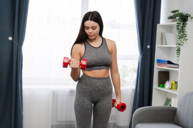 Formda, şık spor giysileri giyen, modern oturma odasında dambıllı, düşünceli, güzel bir genç kız. Spor konsepti