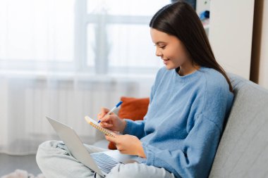 Mutlu gülümseyen genç kız dizüstü bilgisayarını kullanarak not alıyor oturma odasındaki rahat koltukta oturuyor. Çevrimiçi eğitim kavramı