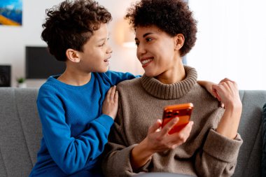 Anne ve oğul kanepede kaynaşıyor, akıllı telefon kullanarak birbirlerine bakıyorlar.