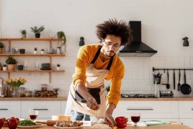 Önlük ve sarı kazak giyen genç adam yemek pişiriyor, parlak bir mutfakta malzemeler serpiyor.