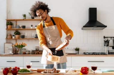 Önlük giymiş genç bir adam, açık bir evde yemek pişirirken yemek malzemeleri hazırlıyor.