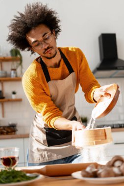 Önlüklü bir adam un seçiyor, parlak bir mutfakta pişirmek için malzemeler hazırlıyor.
