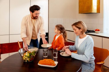 Gülümseyen mutlu aile, baba, anne ve küçük tatlı kızın portresi masada oturuyor, yemek yiyor, evde öğle yemeği yiyor. Yiyecek konsepti