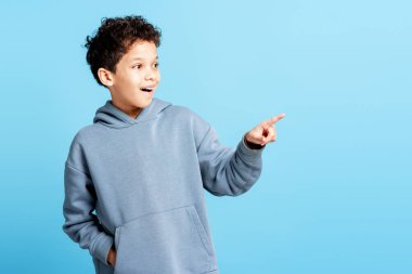 Yakışıklı, gülümseyen küçük çocuk mavi arka planda izole edilmiş fotokopi alanını işaret ediyor. Reklam kavramı