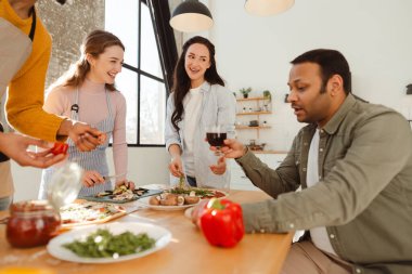Gülümseyen çok uluslu kadınlar ve erkekler birlikte yemek pişiriyor, modern mutfakta pizza yapıyorlar. Arkadaşlık kavramı