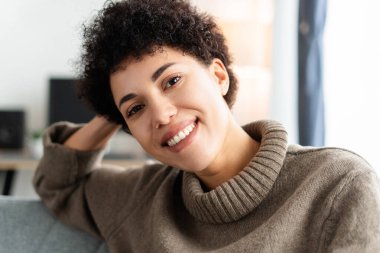 Neşeli, şık günlük kıyafetler giyen güzel bir kadın evde, oturma odasında kameraya bakıyor. Başarılı bir iş kadını pozu. Reklam kavramı