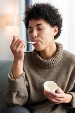 Neşeli, kıvırcık saçlı, evde kapalı gözlerle yoğurt yiyen güzel bir kadın. Beslenme sağlık konsepti