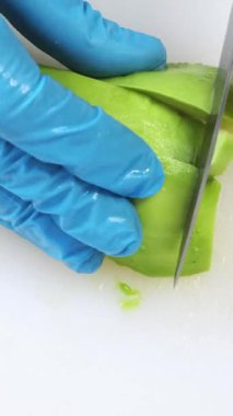 Yeşil avokadonun yarısı beyaz mutfak tahtasında kesiliyor. Mavi eldivenli şef taze sebze doğruyor, demir bıçak. Profesyonel restoranda yemek pişirmek, malzemeleri hazırlamak. Üst alt görünüm