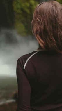 Güçlü bir Bali şelalesinin önünde yemyeşil bir şelaleyle çevrili kendine güvenen bir kadın. Su kayalıklardan aşağı doğru büyüleyici bir güç gösterisiyle akıyor..