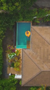 Bir kadının, tropikal lüks bir villadaki yüzme havuzunda yüzen sarı şişme bir yüzükte tatilinin keyfini çıkarışını gösteren hava manzarası.