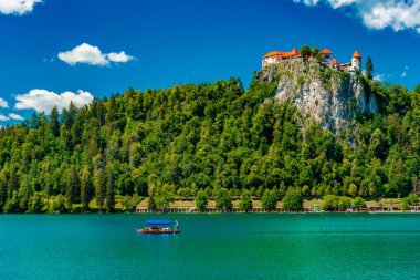 Slovenya 'da Kan Gölü. Yeşil ormanlarla kaplı bir tepede şatosu, turistlerle dolu bir teknesi ve turkuaz suyu olan bir gölü olan resimli bir yaz manzarası.