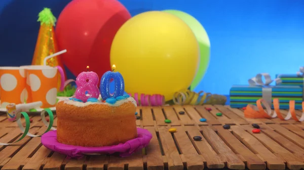 Születésnapi torta gyertyákkal a rusztikus, fából készült asztal háttér színes lufi, ajándékok, műanyag pohár és cukorka a kék falon a háttérben — Stock Fotó