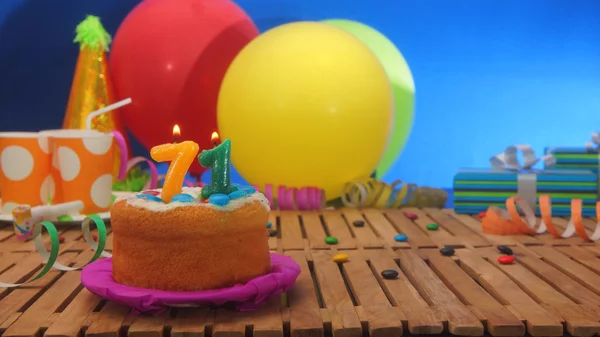 Kue ulang tahun dengan lilin di meja kayu pedesaan dengan latar belakang balon berwarna-warni, hadiah, cangkir plastik dan permen dengan dinding biru di latar belakang — Stok Foto
