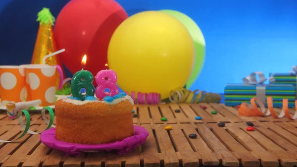 Bolo de aniversário com velas na mesa de madeira rústica com fundo de balões coloridos, presentes, copos de plástico e doces com parede azul no fundo . — Fotografia de Stock