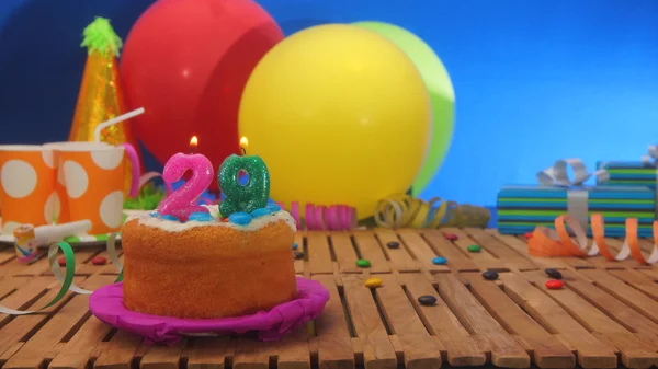 Születésnapi torta gyertyákkal a rusztikus, fából készült asztal háttér színes lufi, ajándékok, műanyag pohár és cukorka a kék falon a háttérben. — Stock Fotó