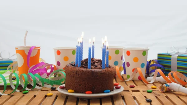 Czekoladowy tort z niebieski palące na rustykalne drewniany stół z tłem kolorowe serpentyny, prezenty, plastikowych kubków z cukierki i białe ściany w tle — Zdjęcie stockowe