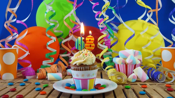 Birthday cupcake with candles burning on rustic wooden table with background of colorful balloons, plastic cups and candies with blue wall in the background — Stock Photo, Image