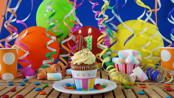 Birthday cupcake with candles burning on rustic wooden table with background of colorful balloons, plastic cups and candies with blue wall in the background