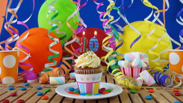 Birthday cupcake with candles burning on rustic wooden table with background of colorful balloons, plastic cups and candies with blue wall in the background