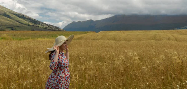 幸せな若いです女性で花のドレス保持彼女の帽子とともに彼女の手だけで歩くの真ん中の小麦畑で曇りの日に青い空と背景の山 — ストック写真