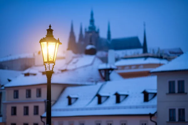 Romantic scene from winter Prague, with the lit lamp during dusk and snow covered roofs in the background, together with the silhouette of the Prague Castle at the horizon.