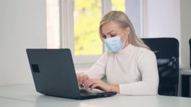 Koruyucu maskeli beyaz bir kadın üniversite öğrencisi ev ödevi yaparken dizüstü bilgisayar kullanıyor. Bilgisayarda öğrenim görüyor. Masada oturuyor, uzaktan eğitim kavramı geliştiriyor.