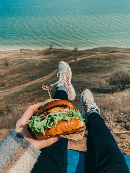 Lezzetli bir fast food sandviçi tutan ve deniz kenarında dinlenen genç bir kadın. Bakış açısı. Deniz kenarındaki hamburgerli kadın görüntüsü.