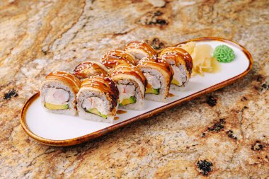 Avokado, yengeç ve yılan balığı gibi taze malzemelerin sergilendiği beyaz bir tabakta suşi ruloları. Ekmekler sosla kaplanmış, wasabi ve zencefille süslenmiş..