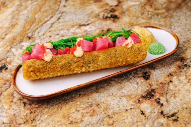 Kurutulmuş ton balığı, avokado ve baharatlı sosla kaplı beyaz bir tabakta canlı bir suşi rulosu sunulur. Tabak turşulu zencefil ve wasabi ile süslenir..